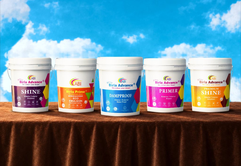 Five ABI paint buckets in a row on a long table draped in chocolate velvet, against a painted sky: Birla Advance Shine, Birla Prime Eco Emulsion, Birla Advance Dampproof, Birla Advance Primer and Birla Advance Premium Shine.